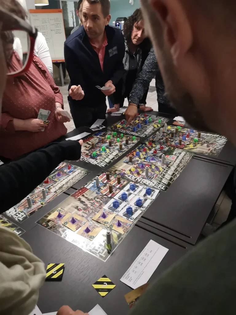 Des participants debout autour d’un grand plateau de jeu couvert de tuiles et de pièces imprimées
