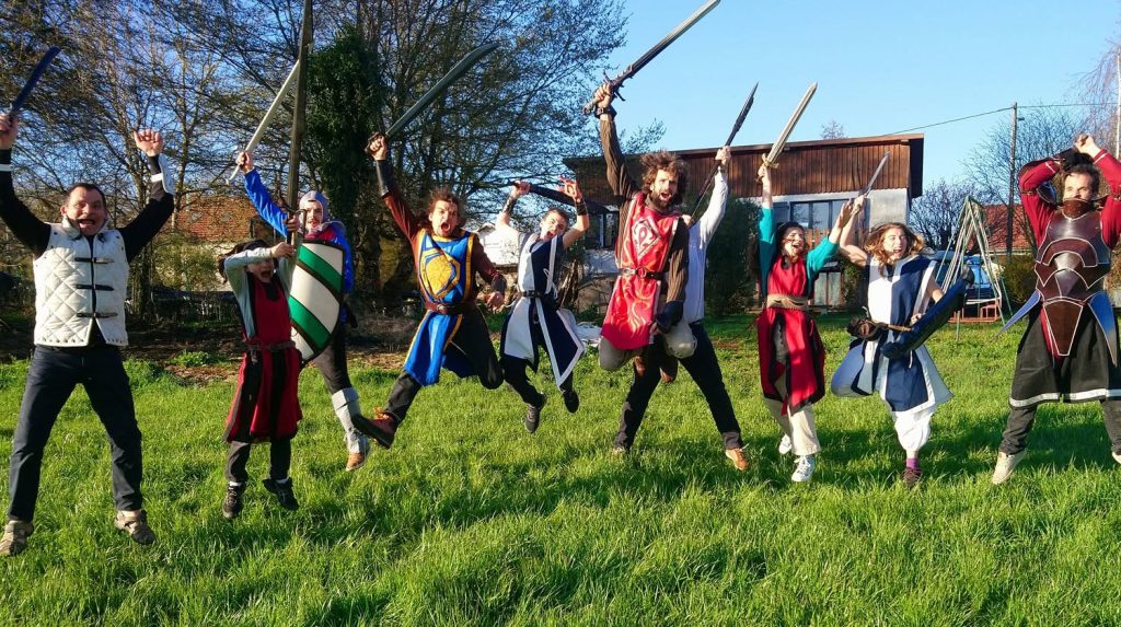 Troll Ball 10Torsions, neuf participants en costume de chevalier sautent en l’air, épées et boucliers en mousse levés, sur une pelouse au soleil